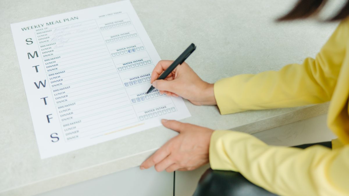 A person writing a weekly meal plan checklist on paper with a black pen